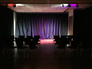 Underground. A stage area with black cloth behind. We see rows of seats with a central aisle.